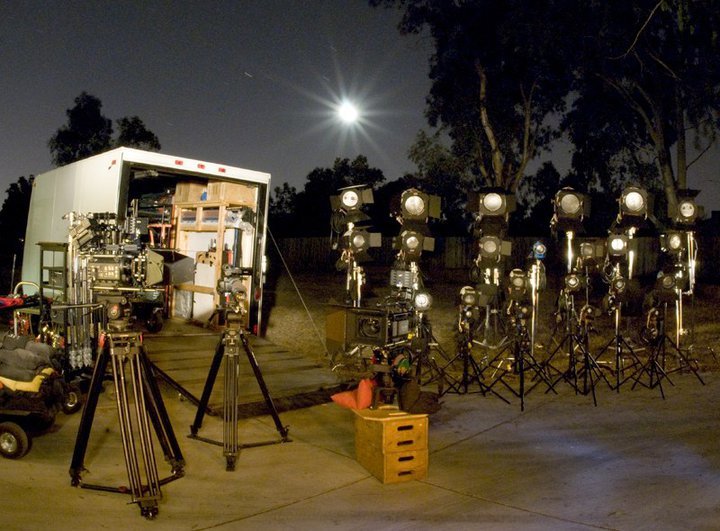 A production truck, cinema cameras and rows of film lights set up at night under a full moon